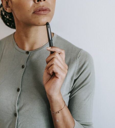 Crop anonymous ethnic female with dark hair in casual outfit touching face with pen and thoughtfully looking away against white wall