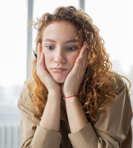 Pensive female student with hands on cheeks looking down while sitting in light room on blurred background while deciding task in frustration