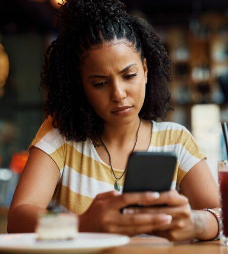 Distraught African American woman reading text message on mobile phone in a cafe.